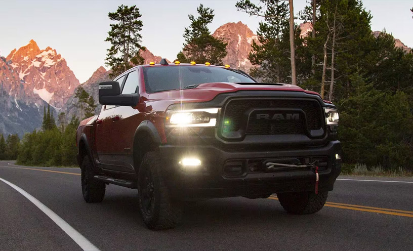 A red 2023 Ram 2500 Rebel Crew Cab with its headlamps on, being driven on a highway with mountains in the background