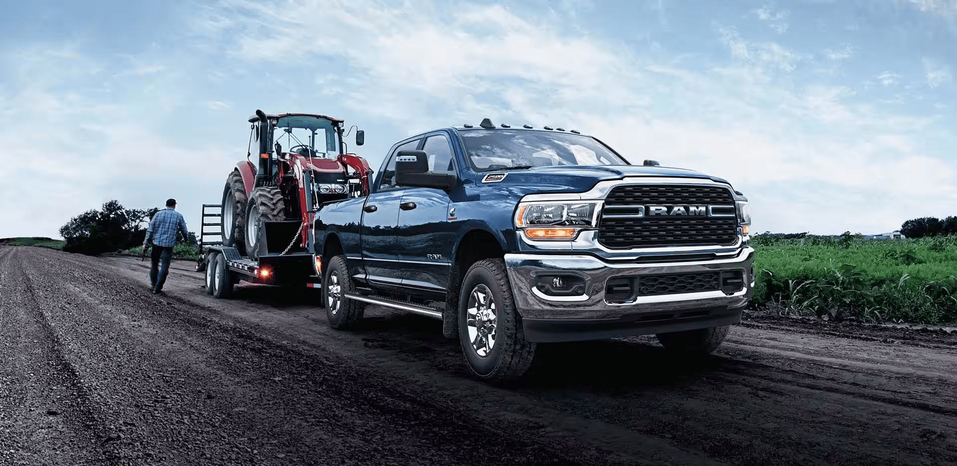 A blue 2024 Ram 2500 Big Horn Crew Cab towing a backhoe on a flatbed trailer.