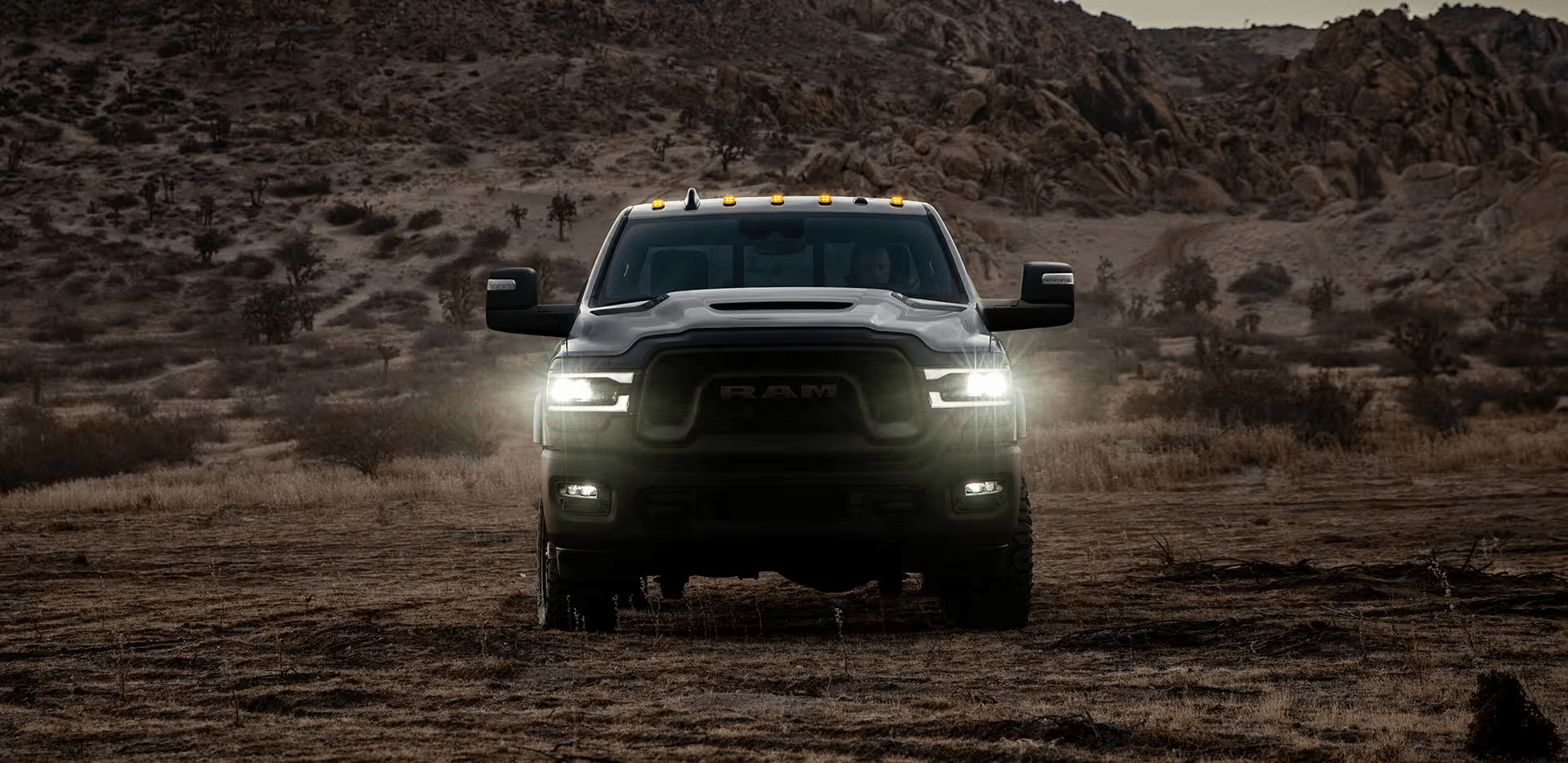 A head-on view of a 2024 Ram 2500 Rebel with its headlamps on, being driven off-road in the desert at dusk.